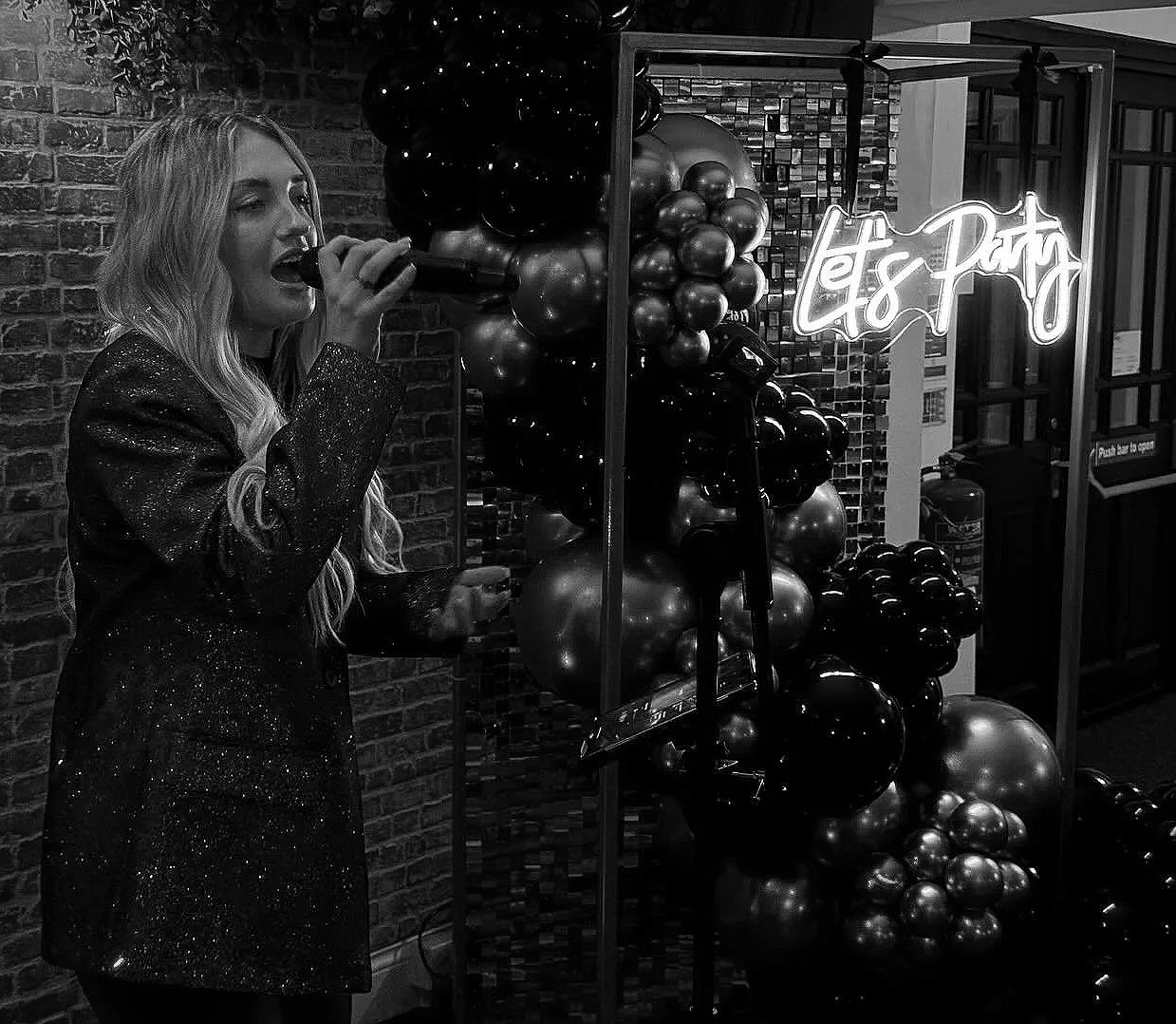 A woman with long wavy hair singing into a microphone at a party, with a neon sign that says 'Let's Party' and decorative balloons in the background.