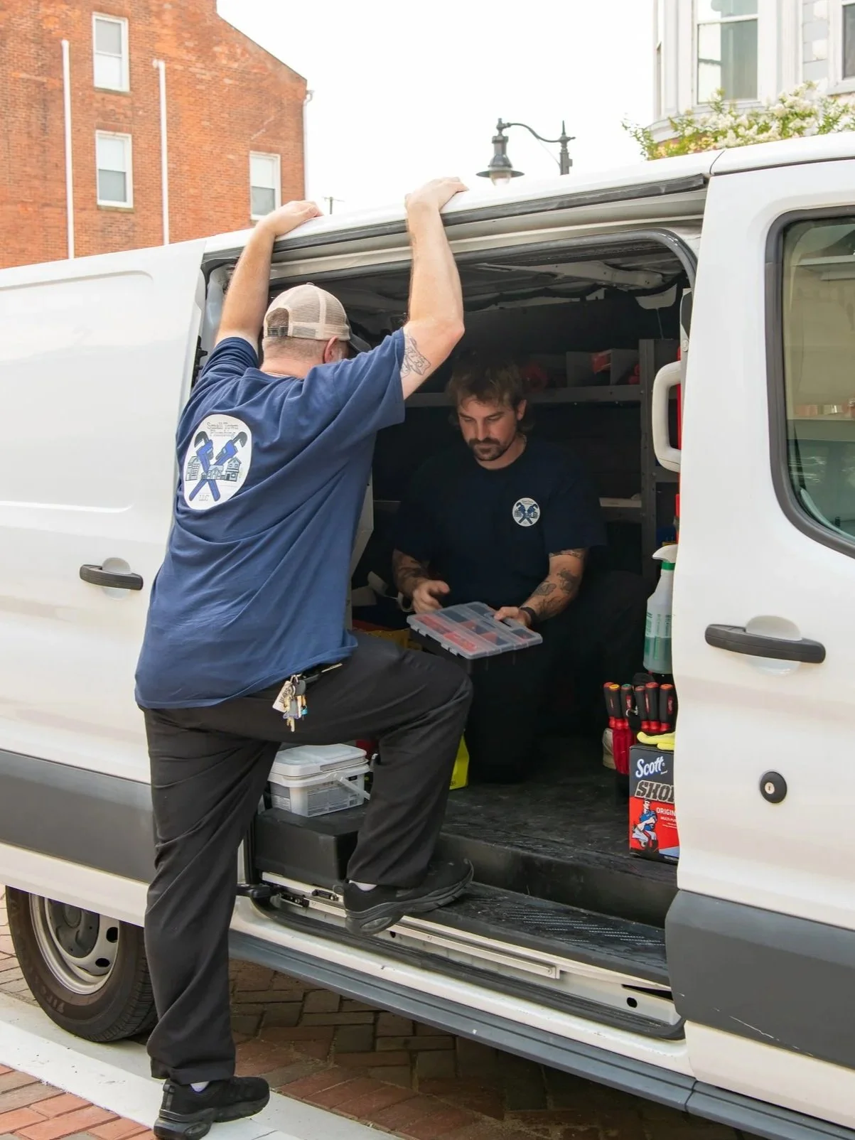 Two men working on a white utility van parked on a street, with brick buildings in the background. One man is standing partially inside the van, and the other is standing outside, holding a toolbox.