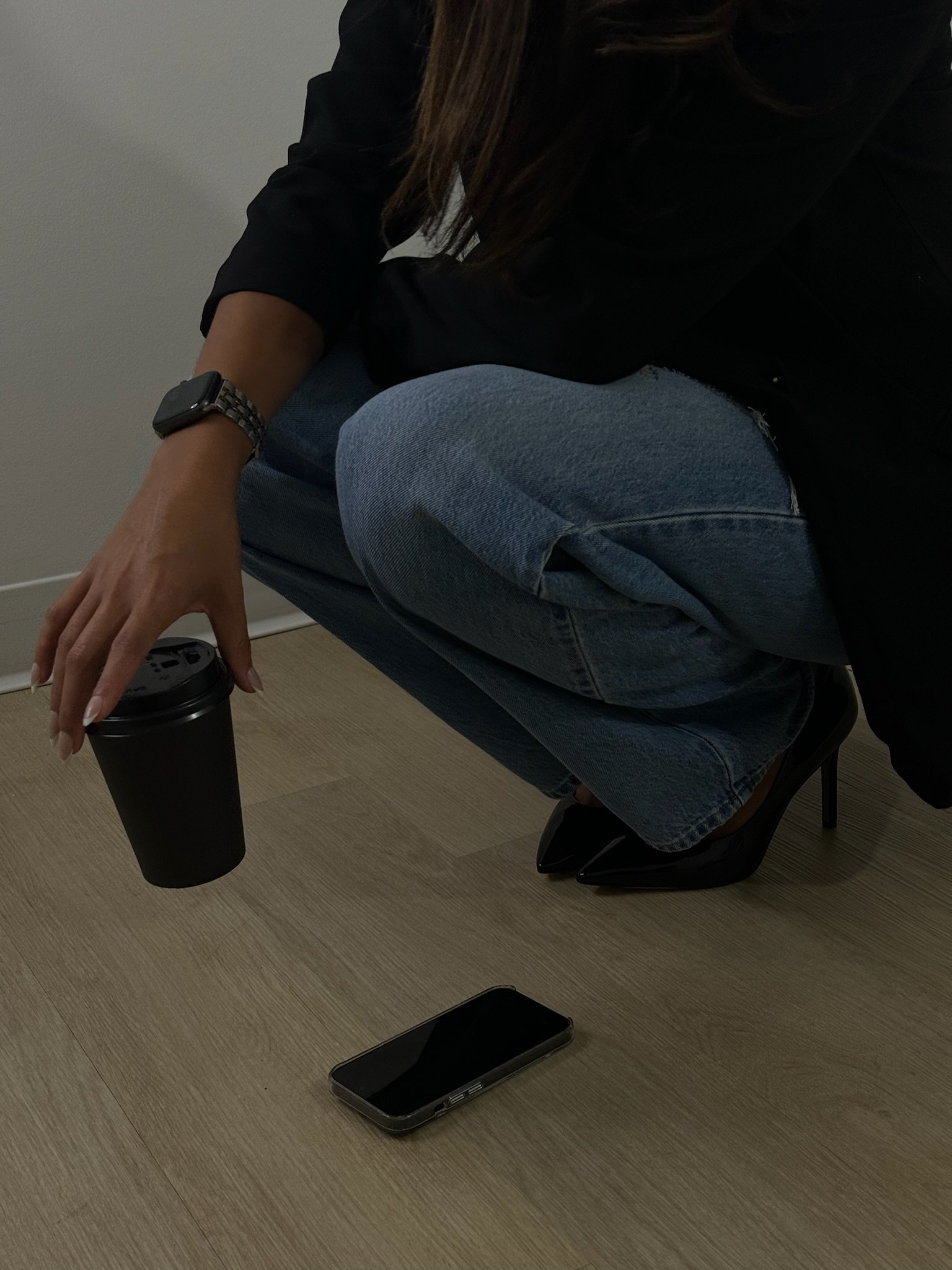 Person squatting near the wall holding a black travel mug, with a smartphone on the wooden floor nearby, wearing a black long-sleeve top, denim jeans, black high heels, and a smartwatch.