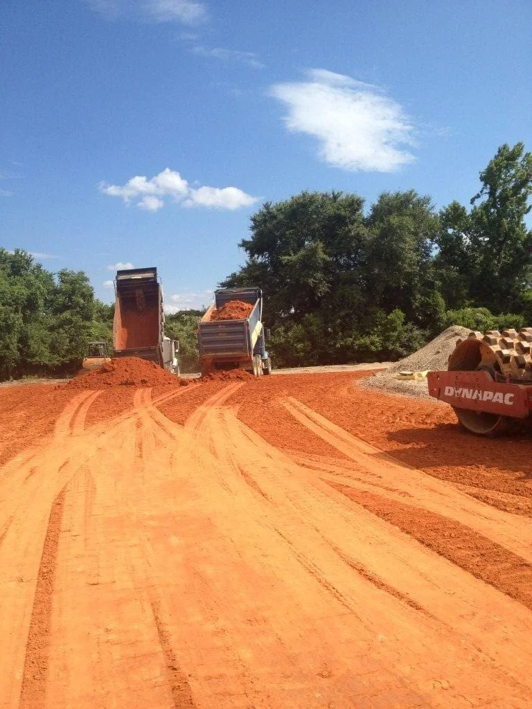 Construction site with two dump trucks unloading dirt onto a red dirt road, a large road roller on the right, green trees in the background, partly cloudy blue sky.
