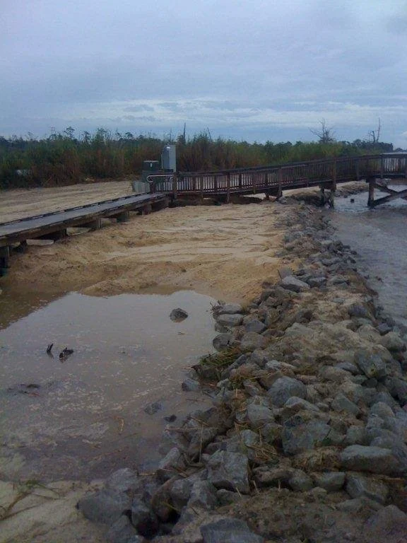 Flooded area with a wooden pier and a sandy pathway, large rocks along the shoreline, overcast sky.