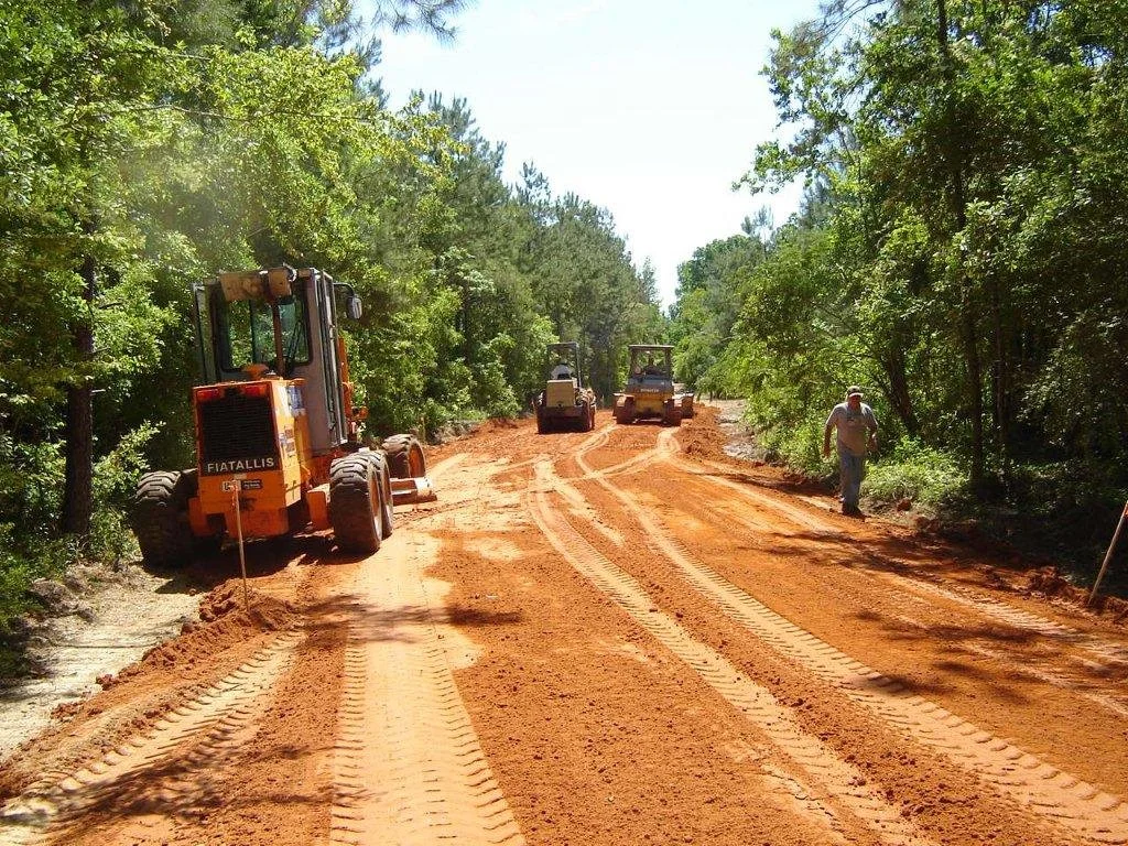 Construction vehicles working on a dirt road surrounded by trees, with a person walking on the right side.