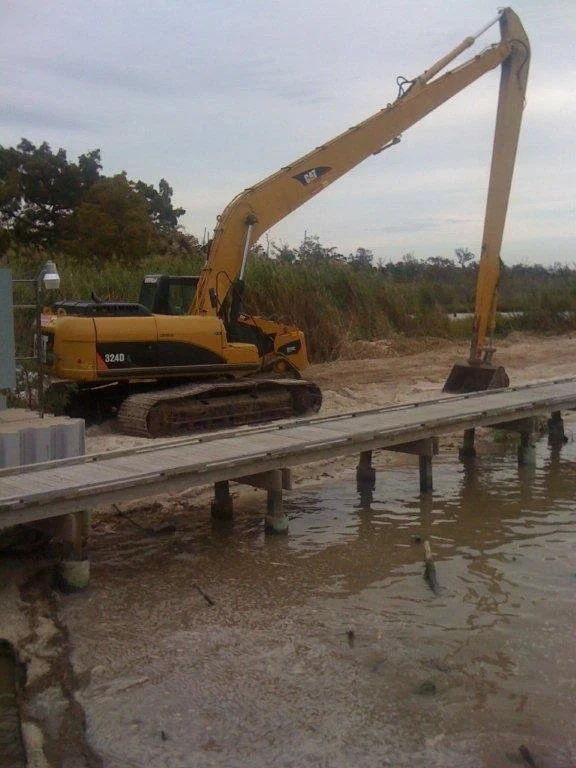 A yellow excavator on a wooden dock over water, with trees and overcast sky in the background.