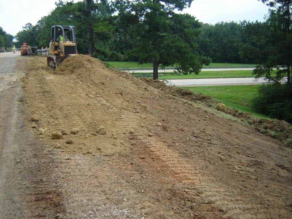 Construction site with dirt and gravel on a road, a small bulldozer working, and trees in the background.