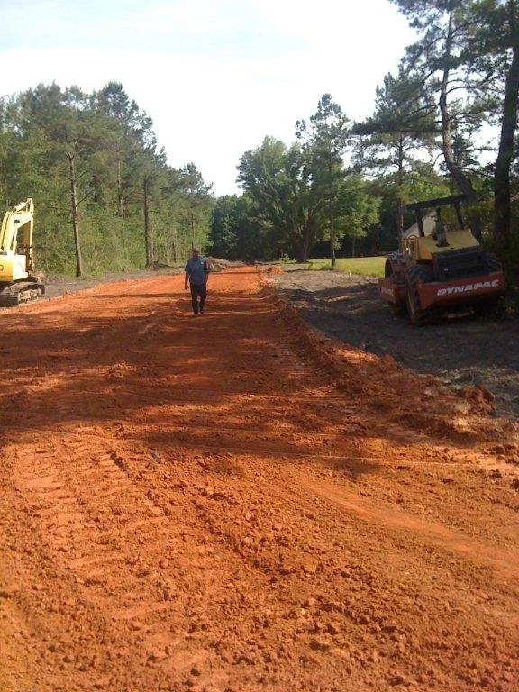 A construction worker walking on a dirt road being prepared or graded, with construction equipment and trees in the background.