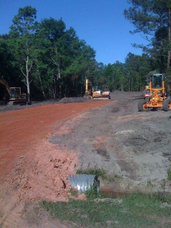 Construction site with dirt and gravel being prepared for paving, with construction vehicles and trees in the background.