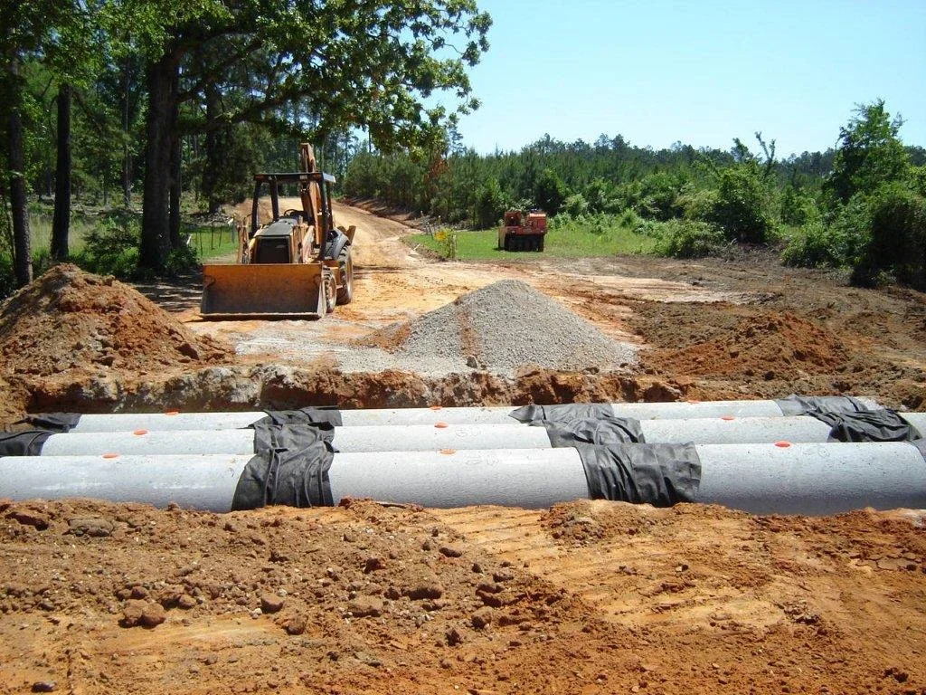 Construction site with pipes laid in the ground, a bulldozer, and gravel mounded on dirt road amid trees.