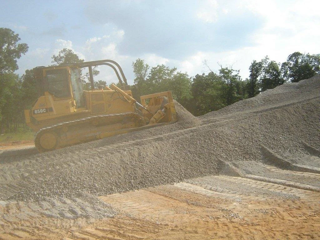 A yellow bulldozer moving a large pile of gray gravel on a construction site with trees and cloudy sky in the background.