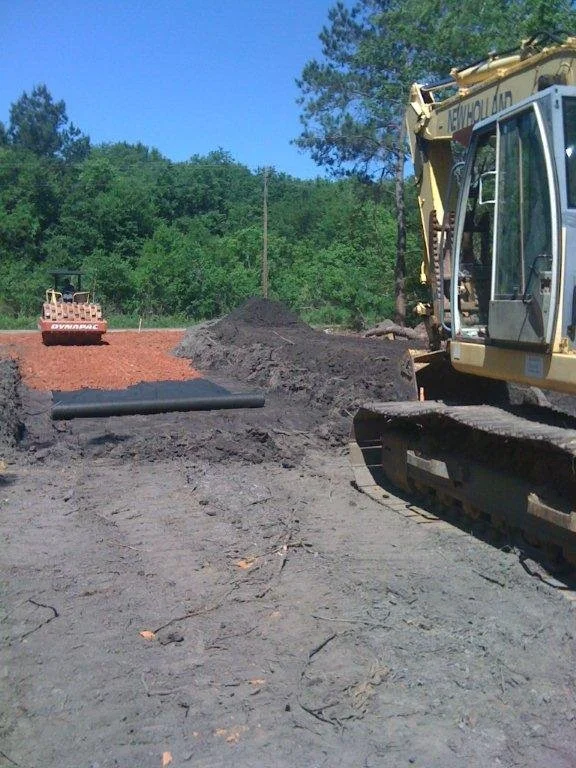 Construction site with equipment and dirt. A bulldozer and a compactor are visible, with piles of dirt and a black pipe on the ground.