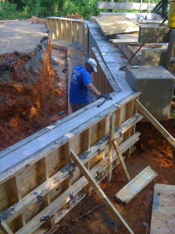 Construction worker in a blue shirt and white hard hat working on a concrete form for a building foundation with wooden supports and framing, surrounded by dirt.