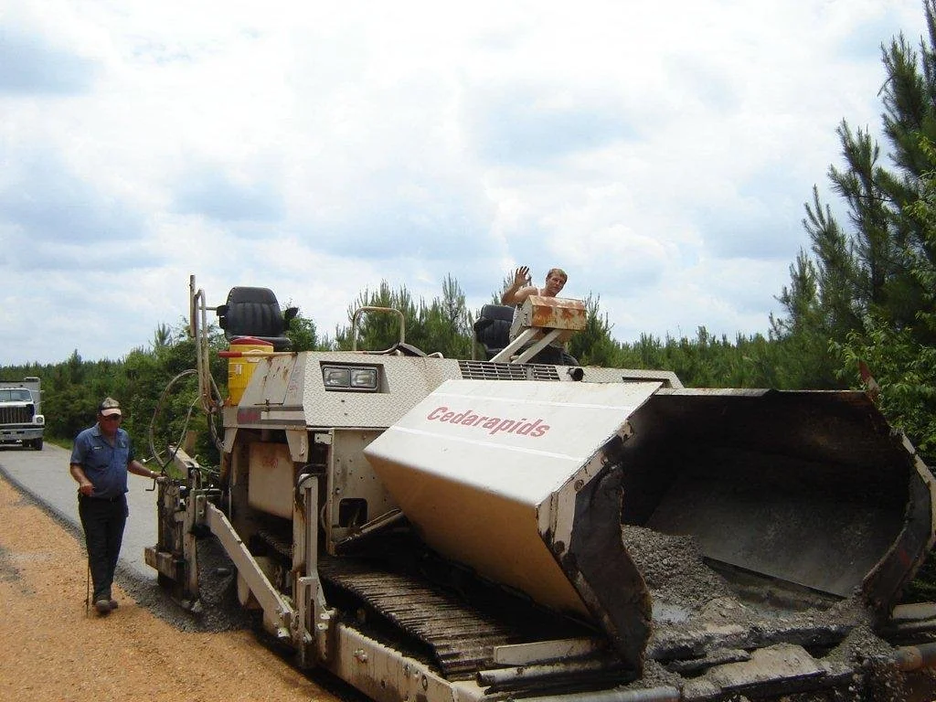 Two men working on a road construction project with a large road milling machine labeled 'Cedarapids' on a partly cloudy day. One man is walking away from the machine, and the other is waving from the top of it.