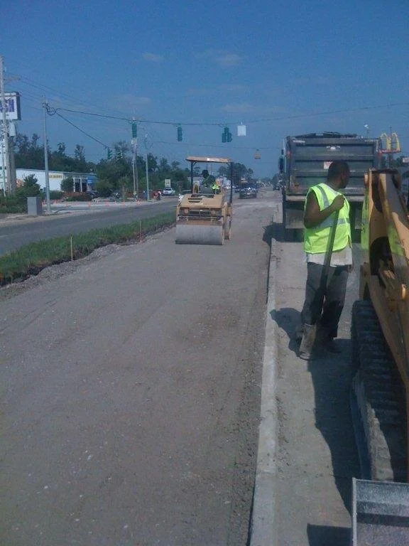 Construction workers operating machinery on a road under a blue sky, with traffic lights and vehicles in the background.
