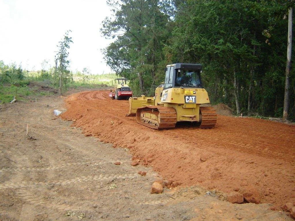 A construction site with two bulldozers leveling a dirt road, with trees in the background.