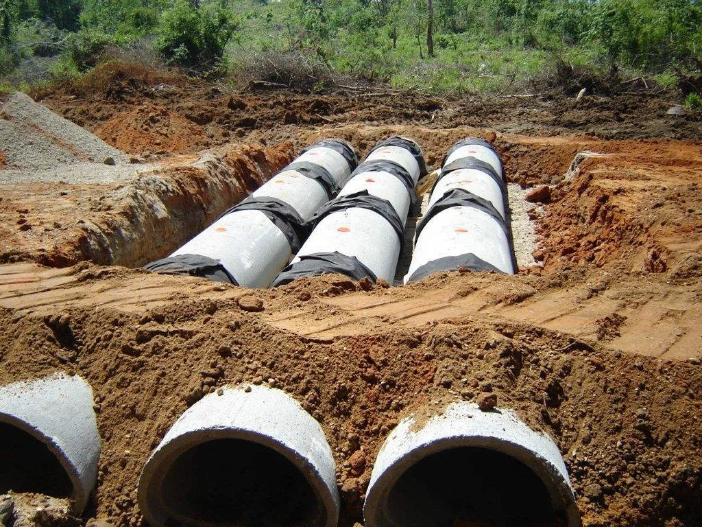 Four large concrete pipes laid in a trench during construction, with surrounding dirt and some green trees in the background.