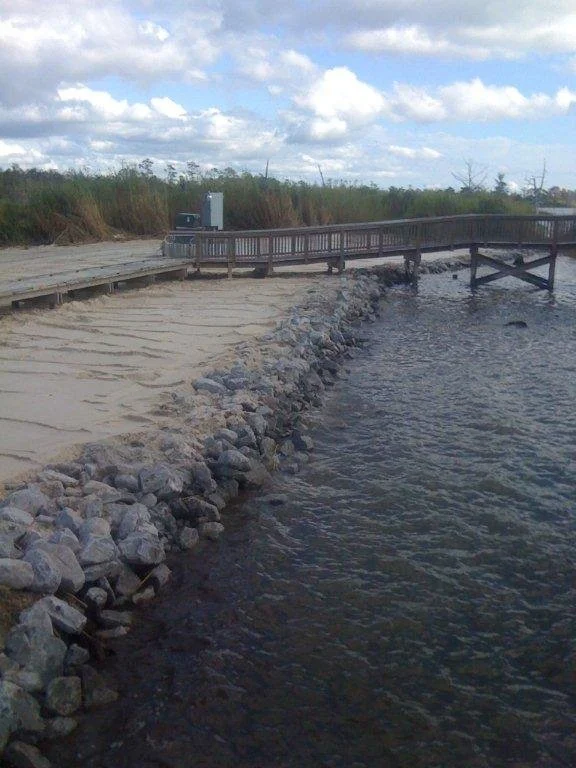 A riverside walkway with a broken part where the wooden platform is collapsed into the water, and a rocky shoreline.