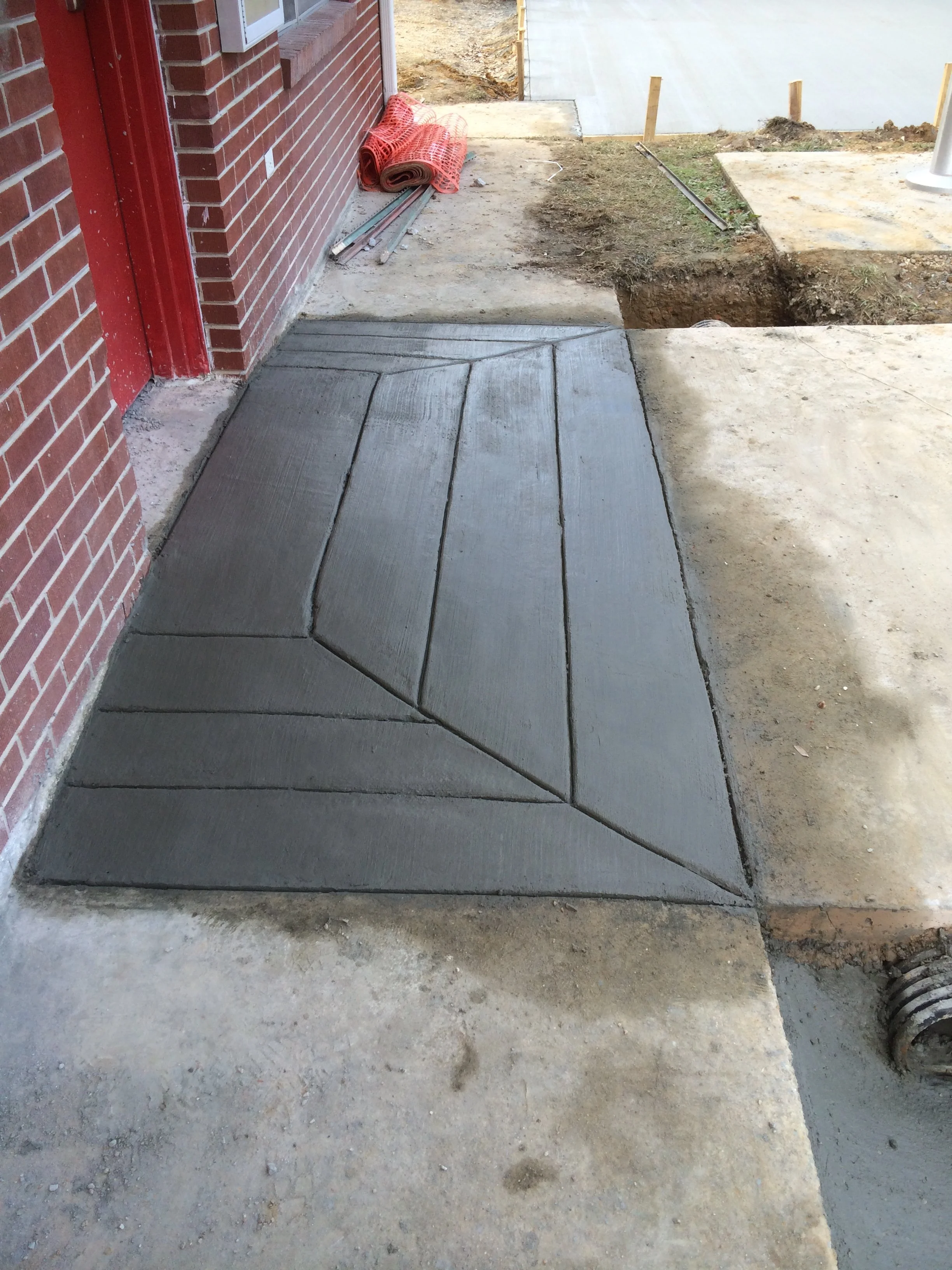 Concrete sidewalk with newly poured sections outside a brick building, with construction materials and a trench nearby.