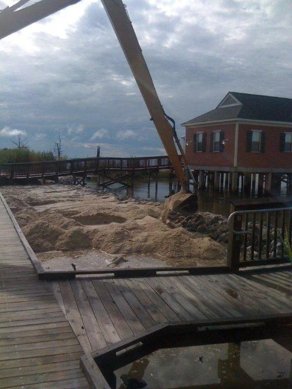 Construction equipment digging soil near a wooden deck and a building on stilts over water