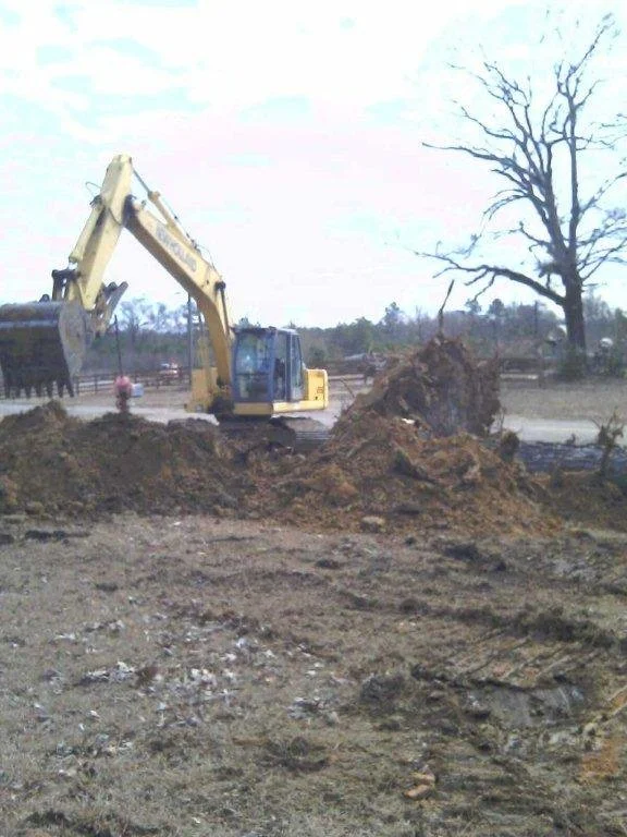 A yellow excavator digging in a construction site with dirt piles, a leafless tree, and a cloudy sky in the background.