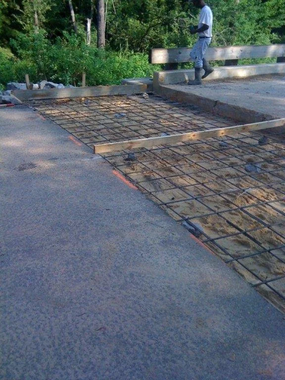Construction worker standing on curb overlooking a concrete slab with rebar, part of road construction in progress, surrounded by wooded area.