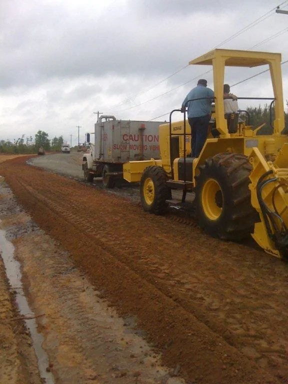 A road construction scene with workers and heavy machinery on a dirt road under cloudy sky.