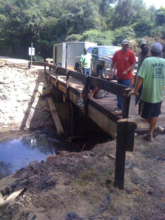 A group of people inspecting a small damaged wooden bridge over a creek on a dirt road, with trucks and trees in the background.