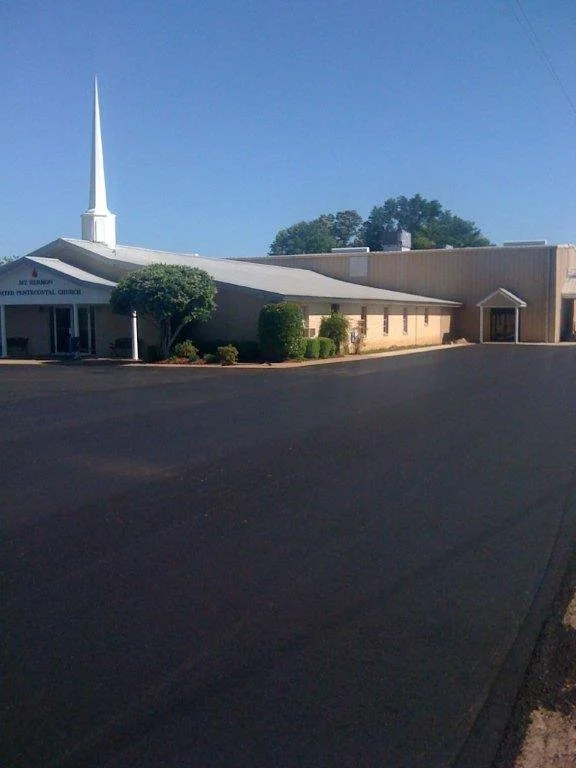 A single-story building with a large white steeple, trees, and shrubs in front, and a black paved parking lot.