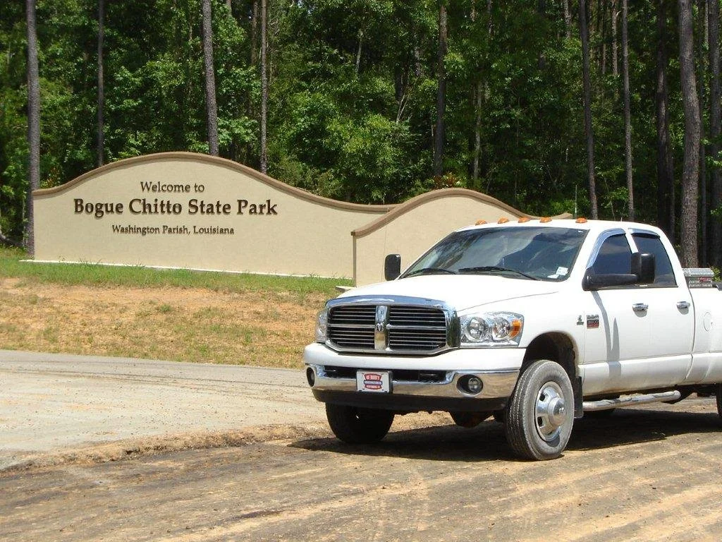 White pickup truck parked in front of a sign for Bogue Chitto State Park in Washington Parish, Louisiana, with a wooded area in the background.