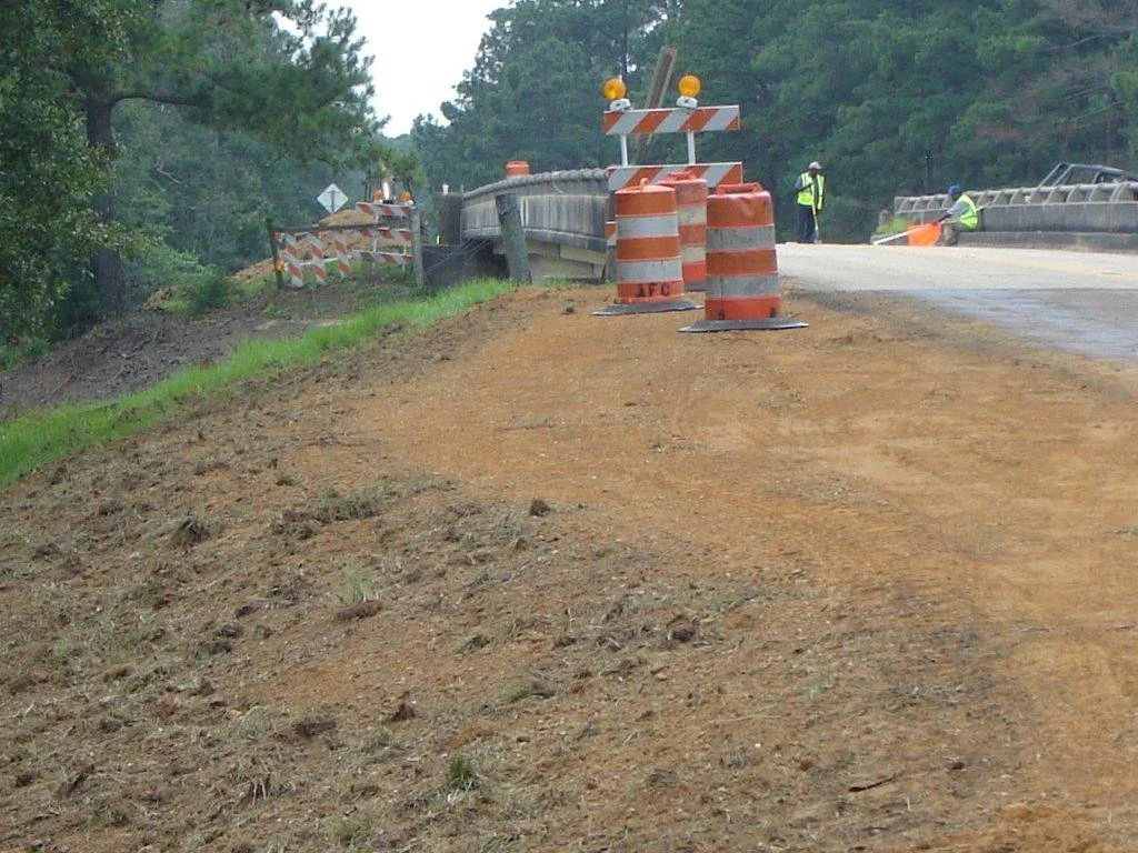 Construction workers repairing a bridge on a road, with orange safety barrels and barriers, and trees in the background.