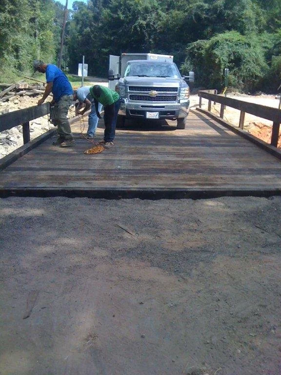 Three workers are on a wooden bridge under construction, with a pickup truck parked behind them. The workers are securing the bridge structure, with one holding a tool and another arranging a chain on the ground.