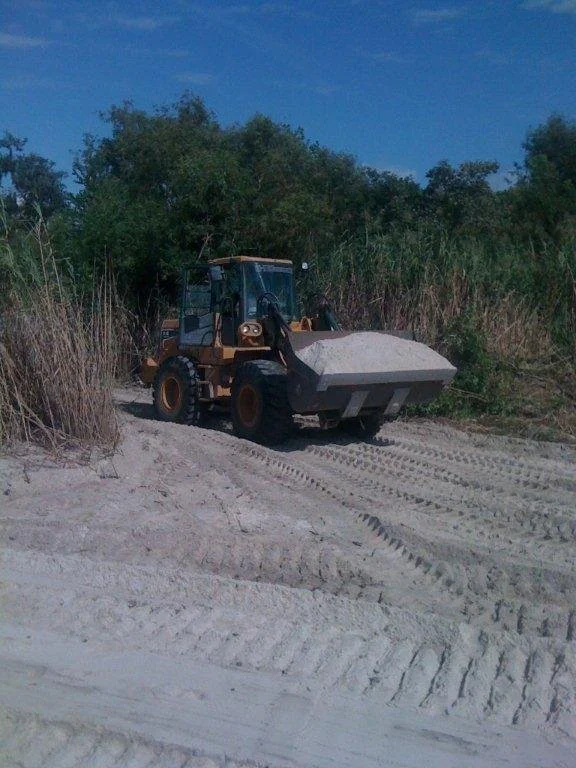 A bulldozer with a scoop full of sand on a dirt path surrounded by tall grass and trees under a clear blue sky.