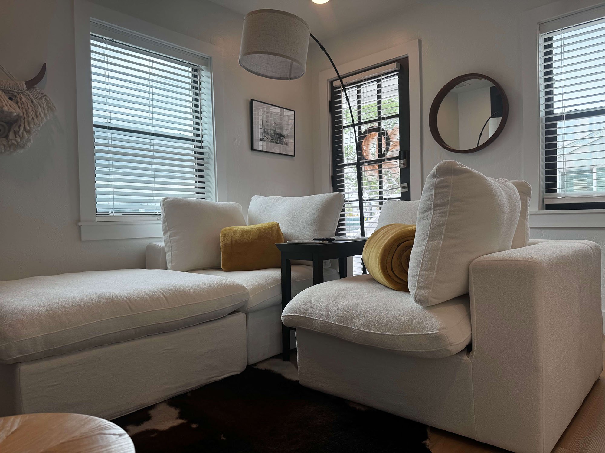 Living room with white sectional sofa, yellow pillow, black side table, and windows with blinds.