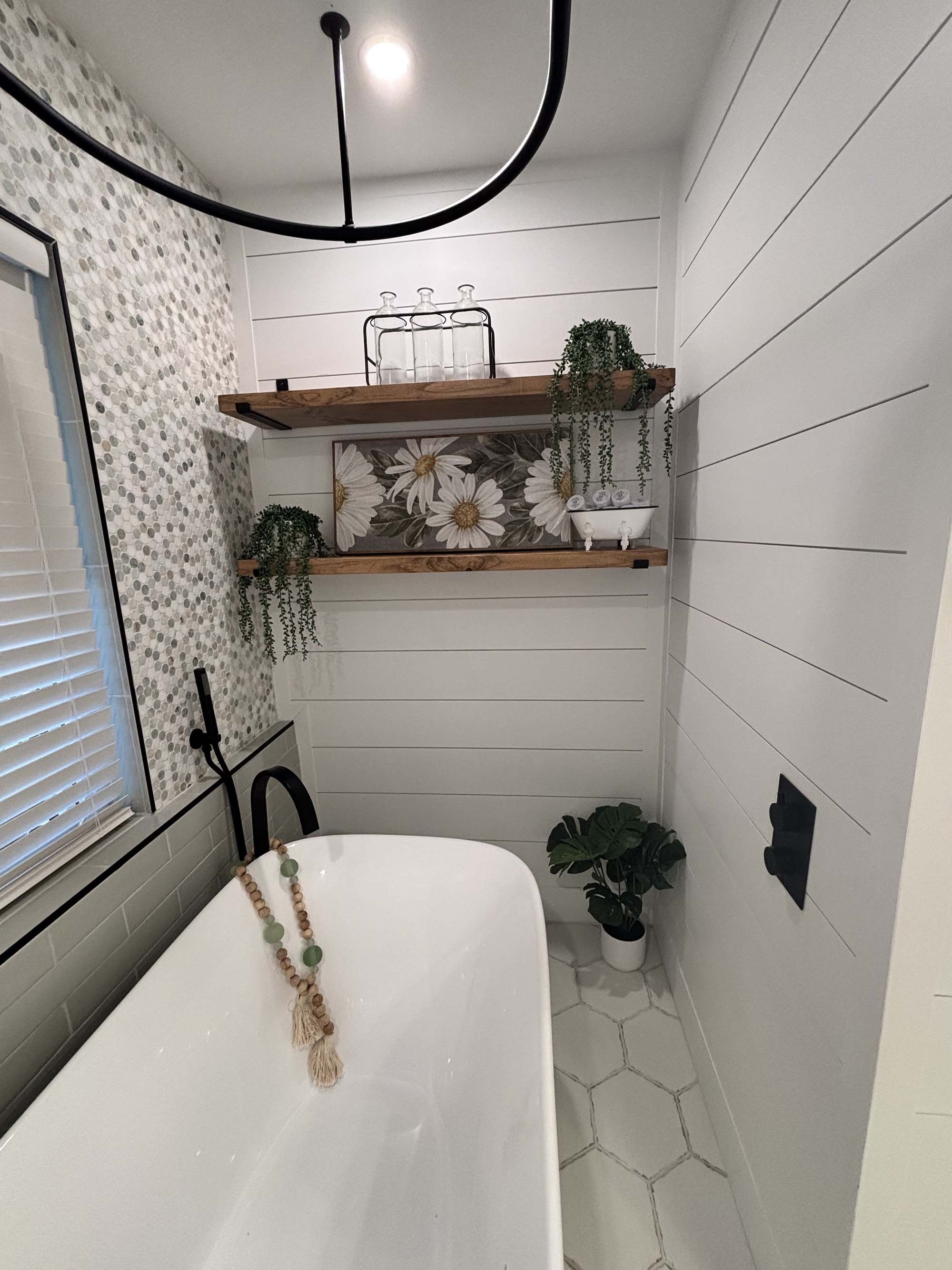 A bathroom with a white bathtub, hexagonal tile floor, white shiplap walls, and two wooden floating shelves decorated with glass bottles, plants, and artwork of daisies. A window with blinds is on the left, and a black faucet is attached to the batht