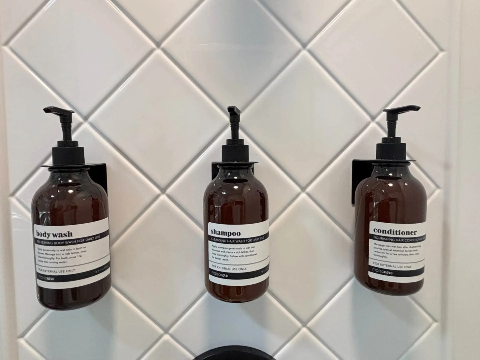Three brown bottles with black pump dispensers mounted on a white tiled bathroom wall. The bottles are labeled 'body wash,' 'shampoo,' and 'conditioner.'