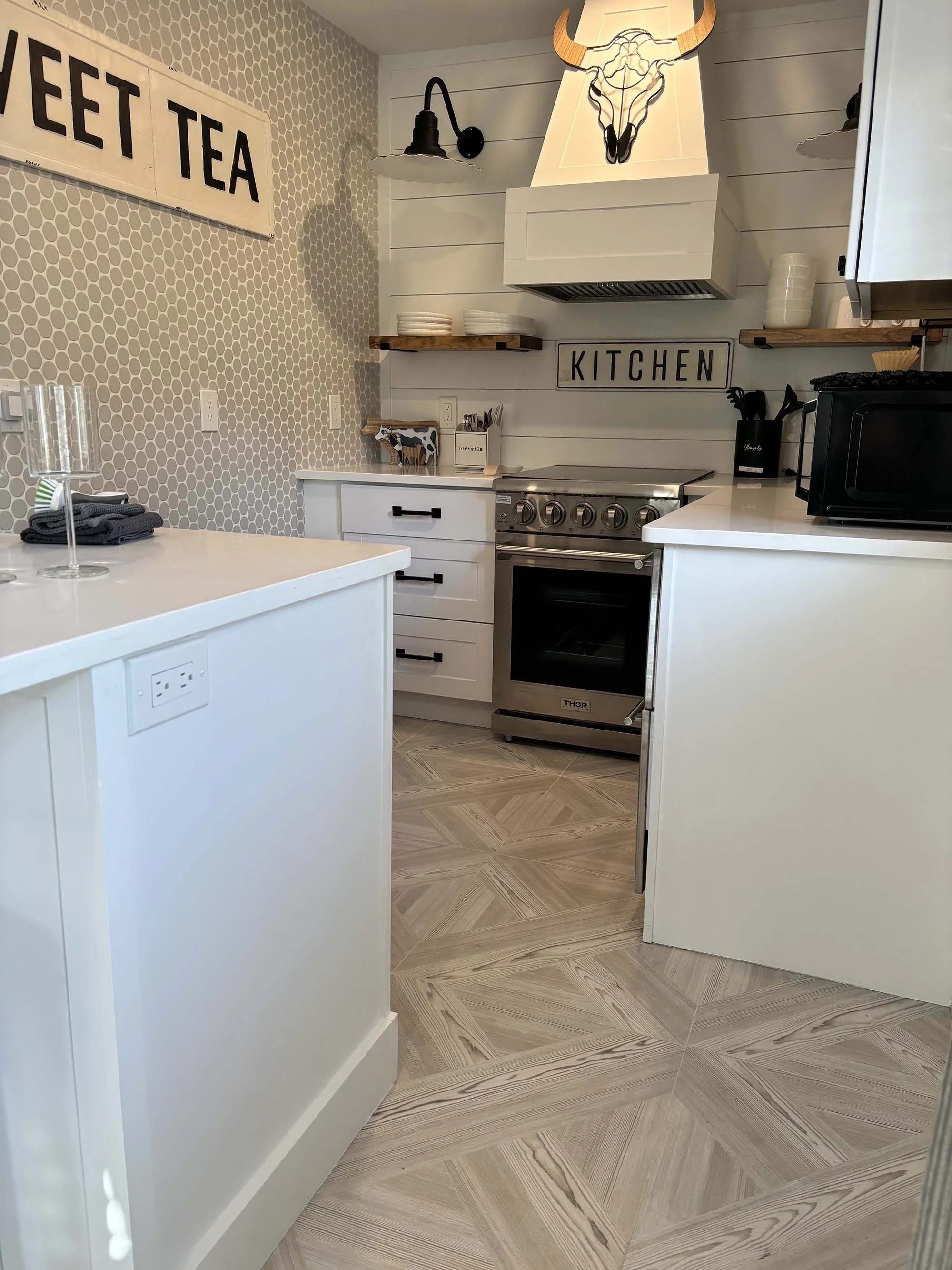 A modern kitchen with white cabinetry, a stainless steel oven, and open wooden shelves on a shiplap wall. Decor includes a mounted bull skull with horns, a black wall-mounted lamp, and a sign reading 'KITCHEN'. There is a black microwave and utensil 