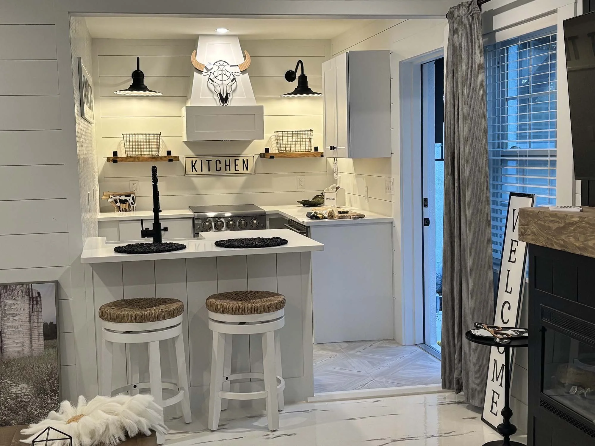 A cozy modern kitchen with white wood-paneled walls, black accents, and farmhouse decor. It features a kitchenette with a stove, a small island with two barstools, a decorative bull's head above the vent hood, and a sign that says 'KITCHEN'. There is