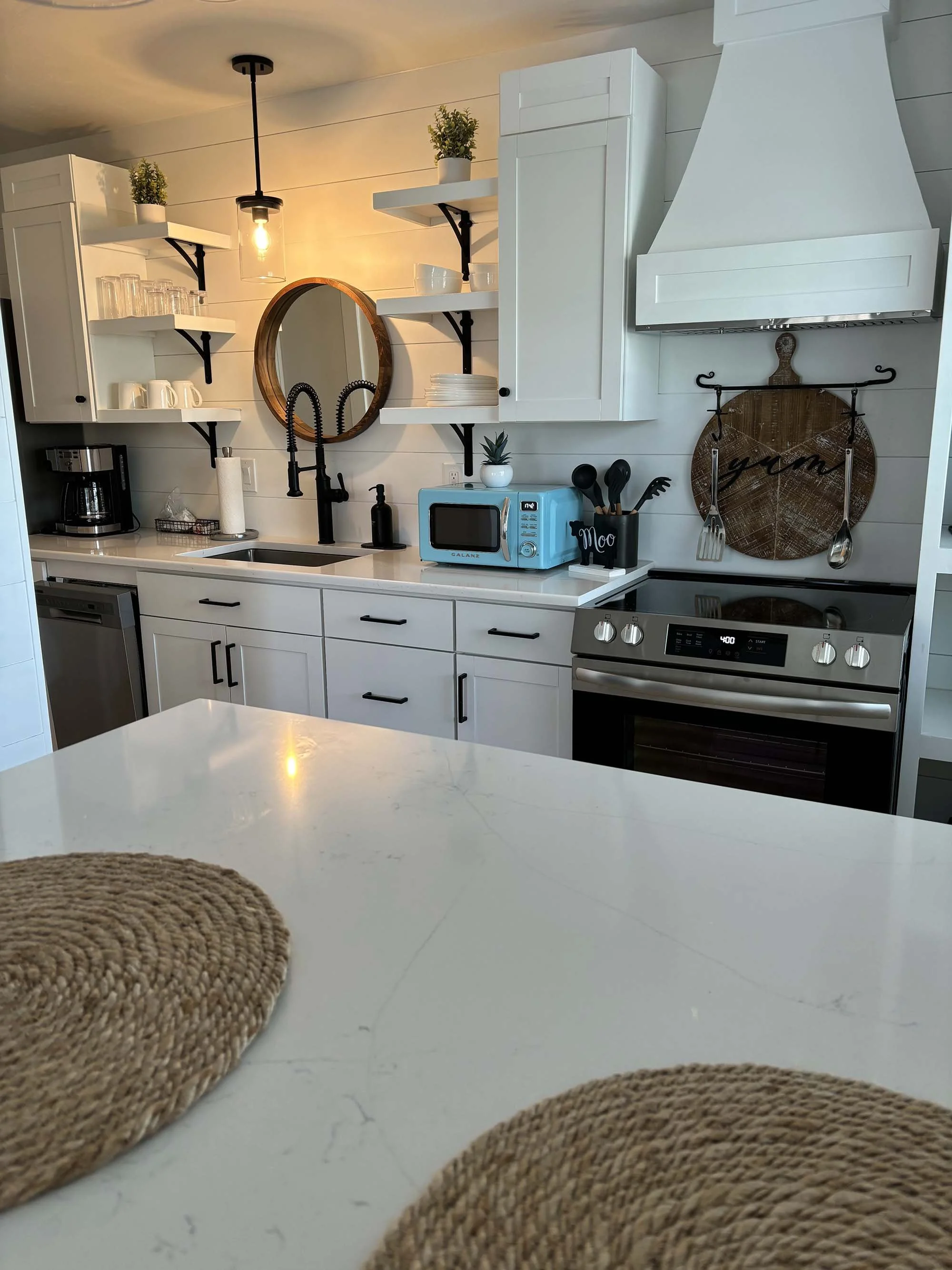Modern kitchen with white cabinets, black hardware, open shelving with dishes and plants, black faucet, blue microwave, black kitchen utensils, and stainless steel stove and dishwasher. Woven placemats on a white countertop in the foreground.