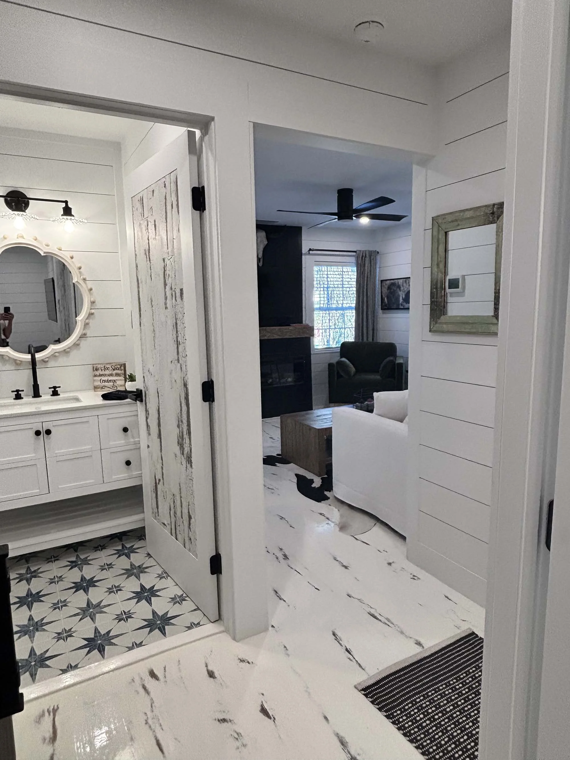 View of a modern interior space with a bathroom on the left and a living room in the background, featuring white walls, marble flooring, a mirror, and furniture.