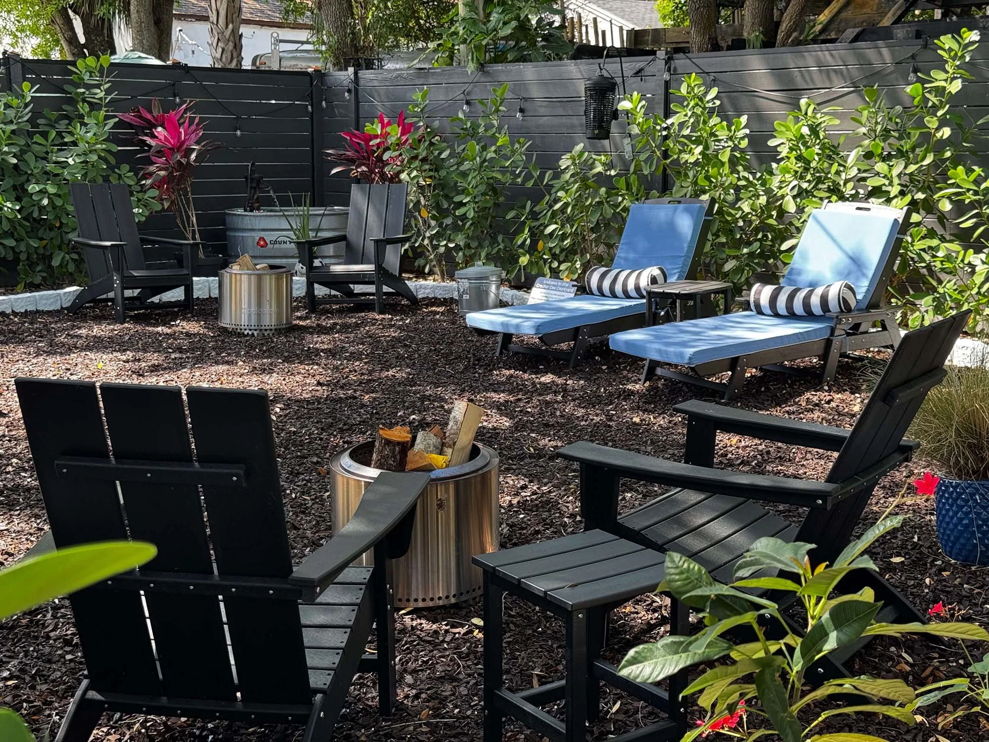 A backyard patio area with black Adirondack chairs, blue lounge chairs with striped pillows, and small side tables, surrounded by green plants and a black fence.
