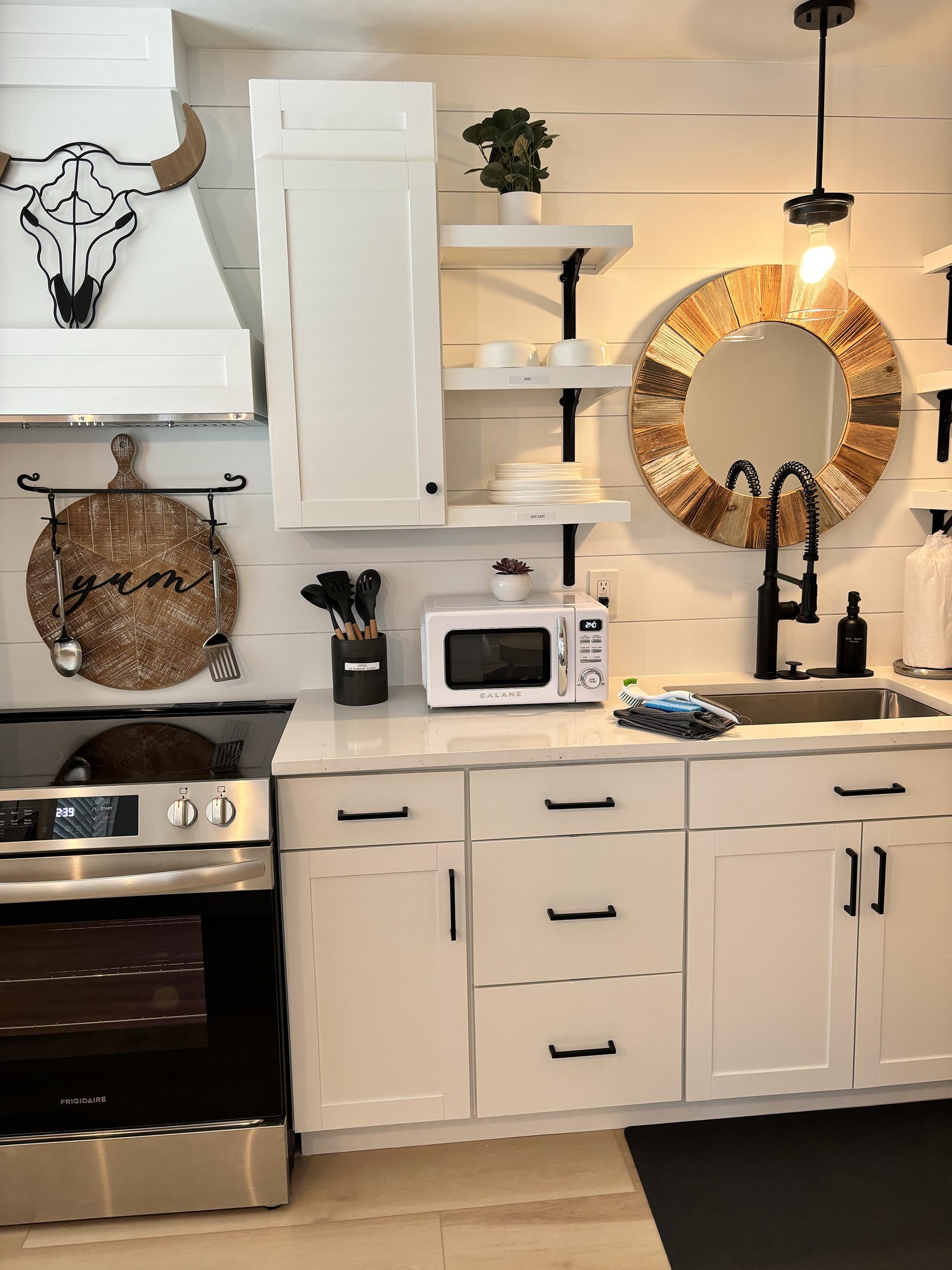 Modern kitchen with white cabinets, black hardware, a stove, microwave, sink, black faucet, round wooden mirror, open shelves with dishes, decorative plant, wall art, and a black hanging light.