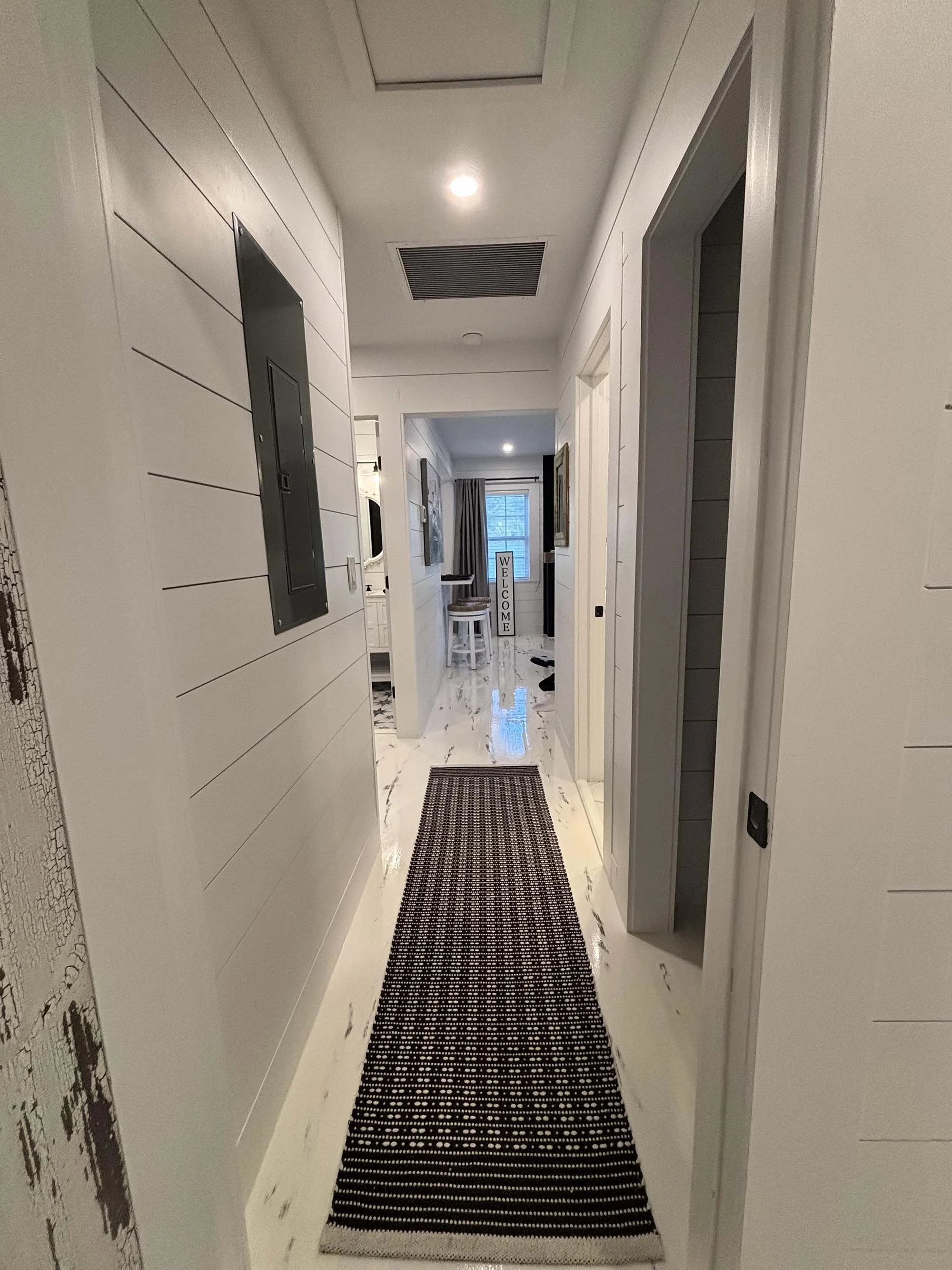 Modern hallway with white shiplap walls, a black and white runner rug, and view into a living space with a welcome sign by the window.