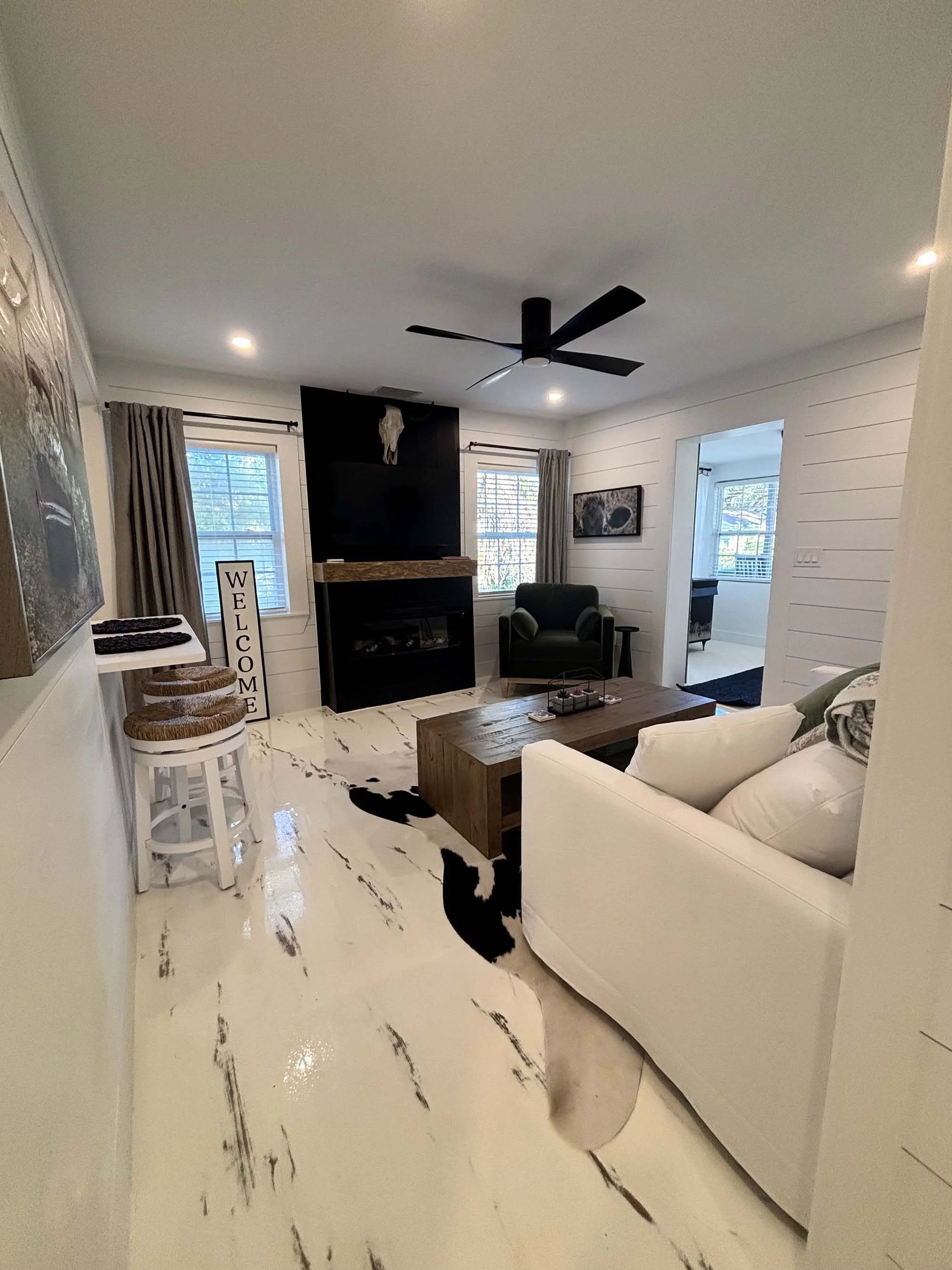 Living room with white walls, black ceiling fan, two windows with beige curtains, brown wooden coffee table, white sofa with pillows, black armchair, and fireplace with a skull decoration above. There are bar stools with woven seats and a welcome sig