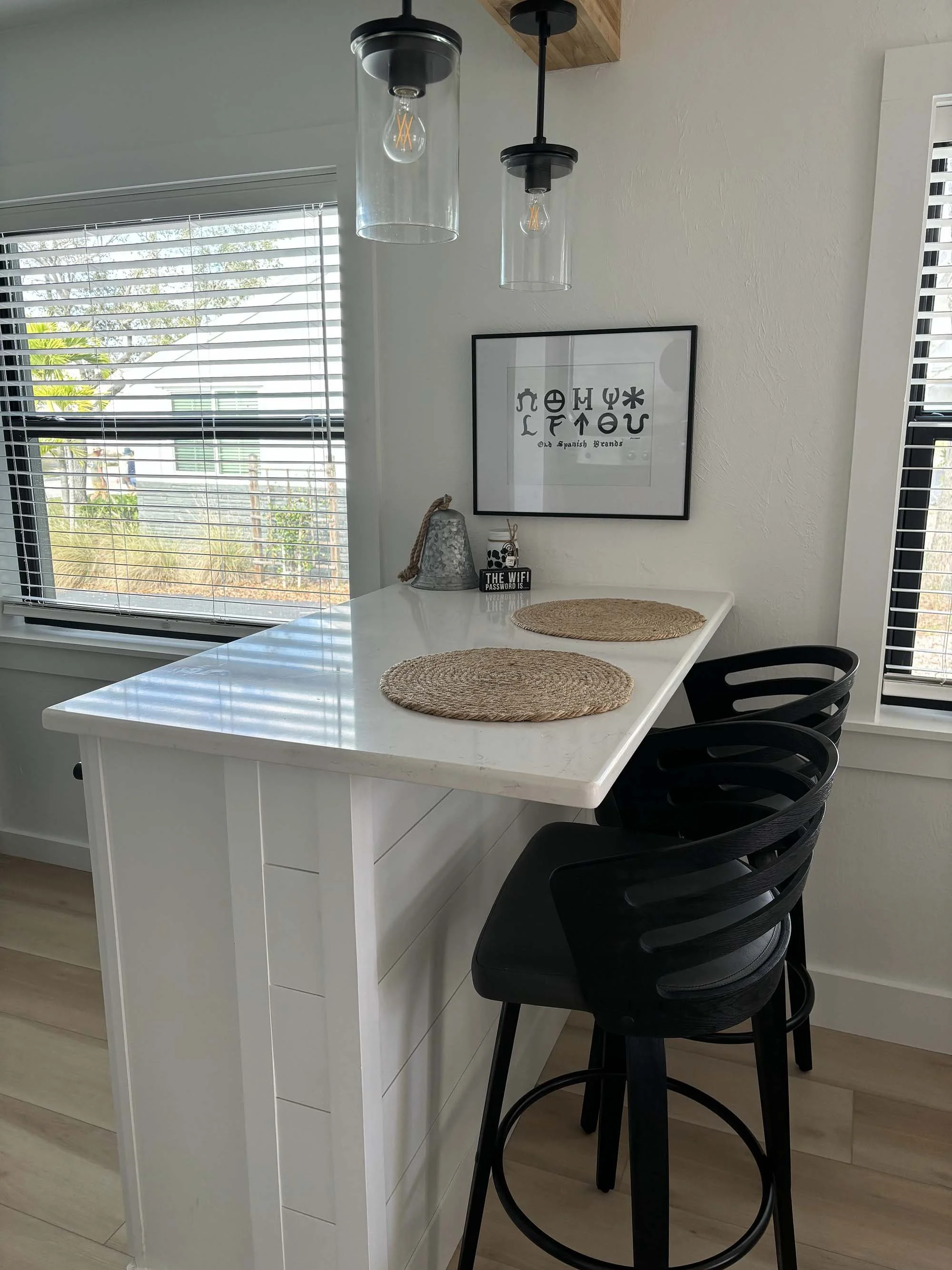 A kitchen island with two black bar stools, woven placemats, and a wall art print of an eye chart with Spanish letters. There are two pendant lights hanging above the island and a window with blinds to the left, showing greenery outside.