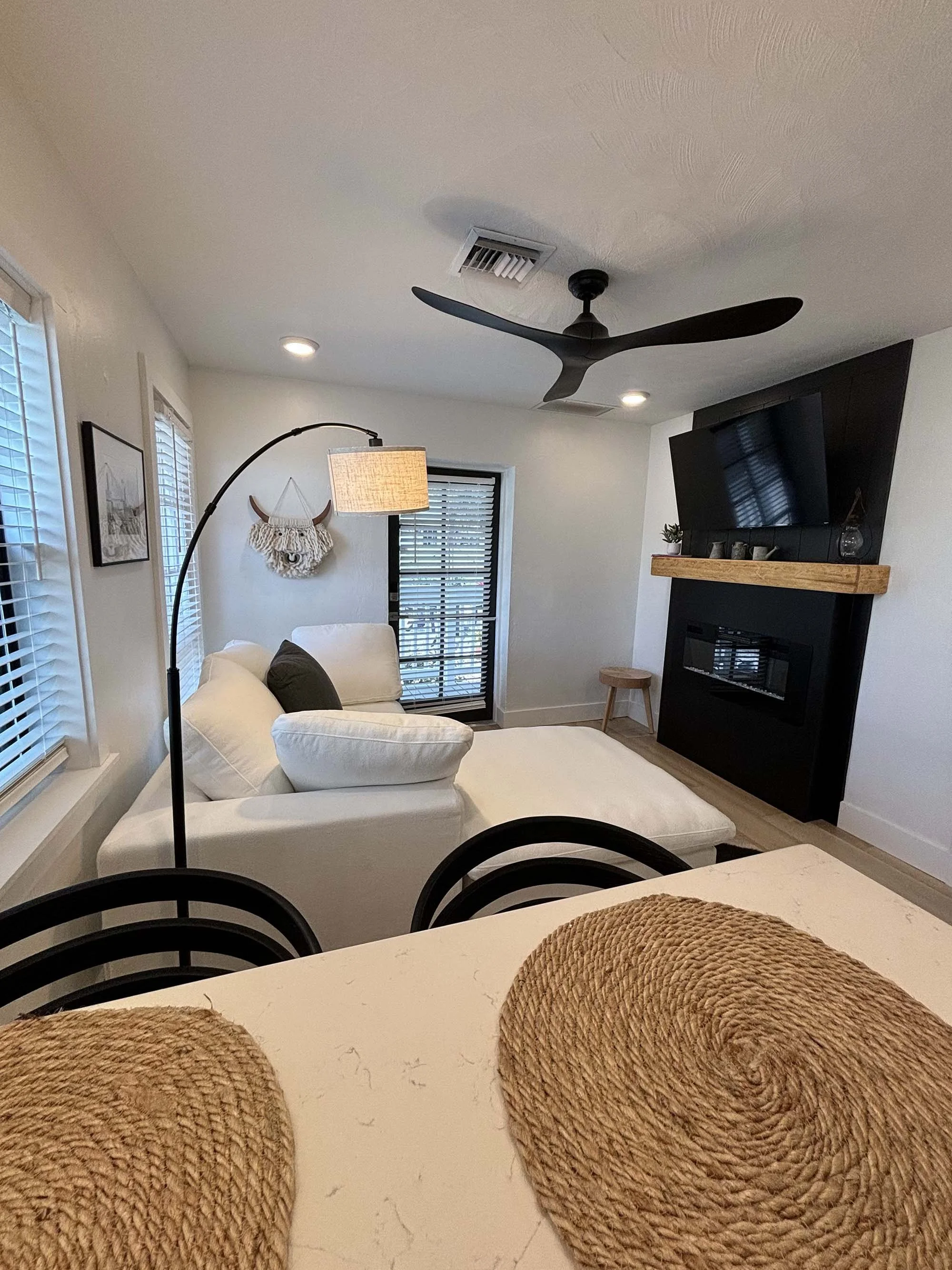 A cozy living room with white walls, a black and wooden fireplace, a large white sofa with pillows, a black standing lamp with a beige shade, a ceiling fan, and windows with blinds. There are decorative items on the mantel and a wall hanging behind t