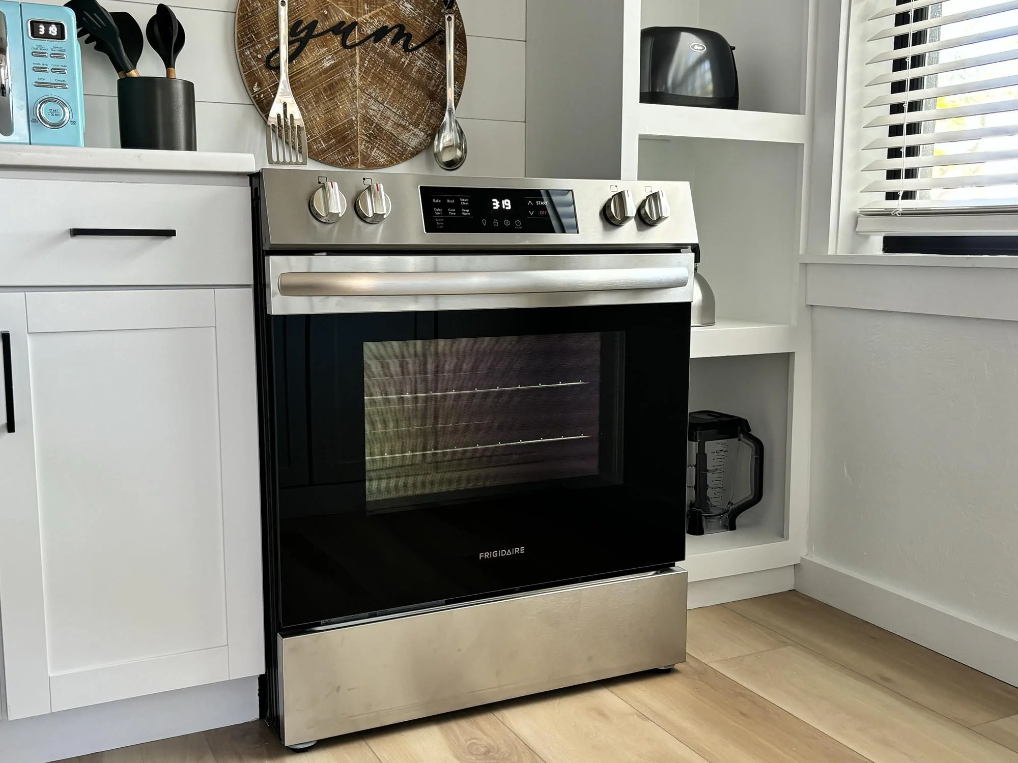Stainless steel and black electric oven with digital display and control knobs, placed in a bright kitchen with white cabinets, wood floors, and a window with blinds.