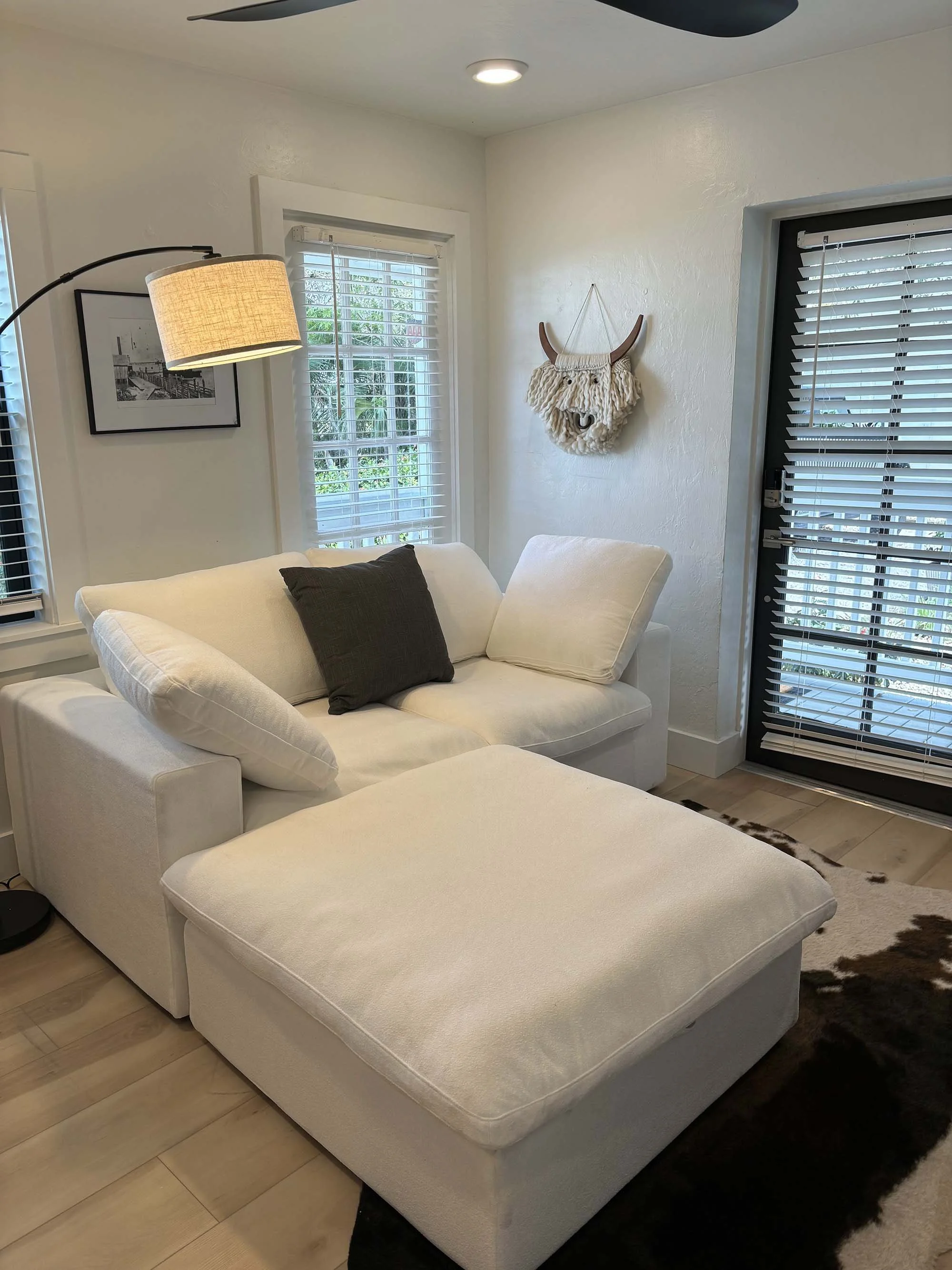 A cozy living room with a white sectional sofa, a black cushion, and a matching ottoman, near windows with white blinds. A modern floor lamp with a beige lampshade, wall art, and a decorative wall hanging resembling a horned animal head are visible.