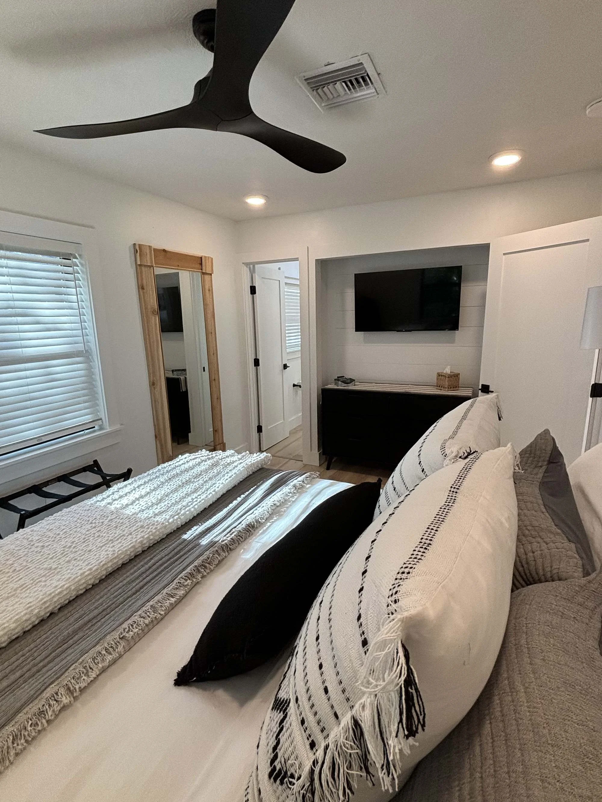 A bedroom with a white bed, black and white pillows, window with blinds, a black ceiling fan, a wall-mounted TV, a mirror with a wooden frame, a black dresser, and a standing lamp.
