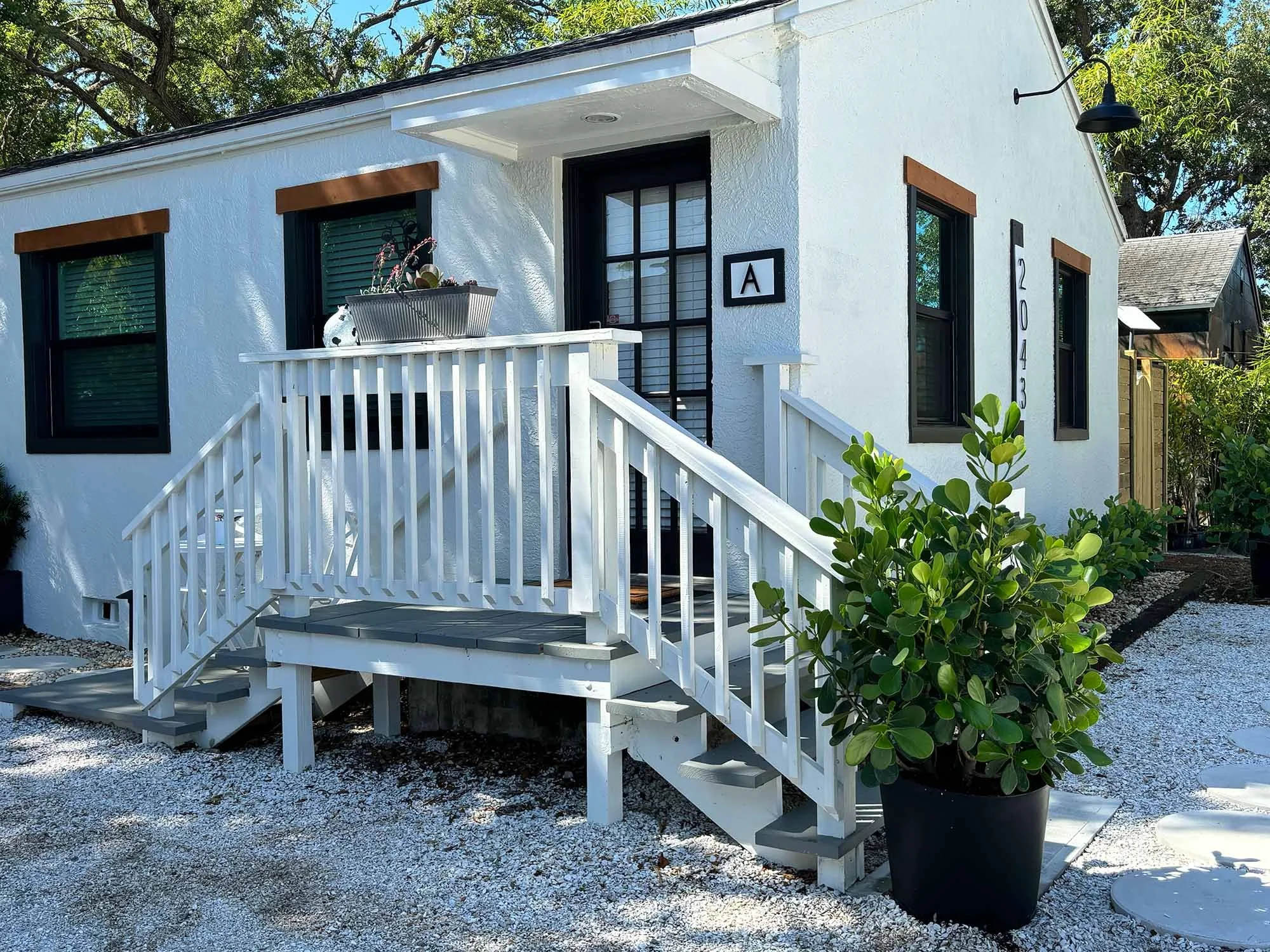 White house with black-trimmed windows, a wooden staircase with white railing, a large potted green shrub, and a gravel pathway.