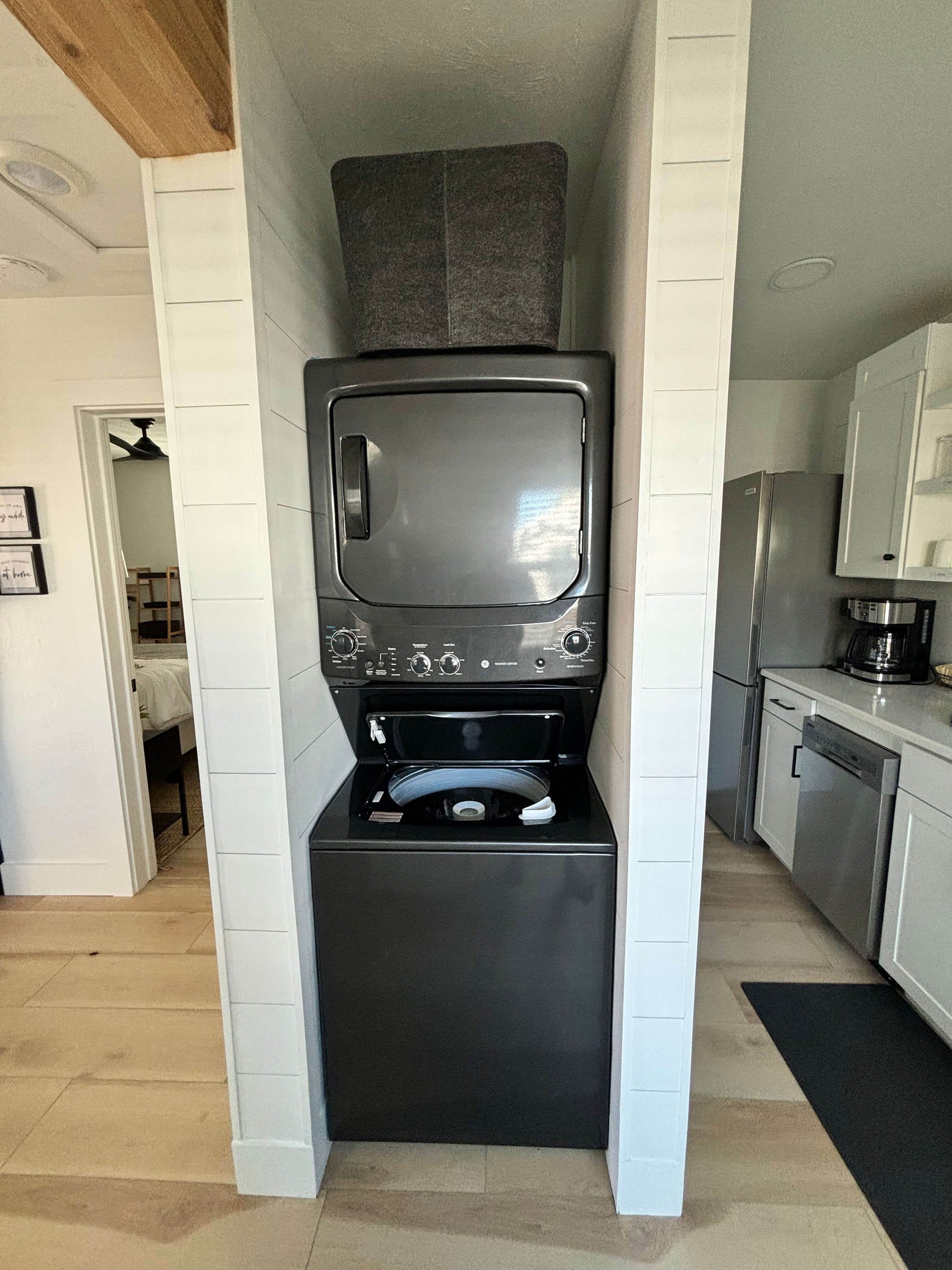 Black stacked washer and dryer in a narrow space in a modern kitchen with wood flooring and white cabinets.