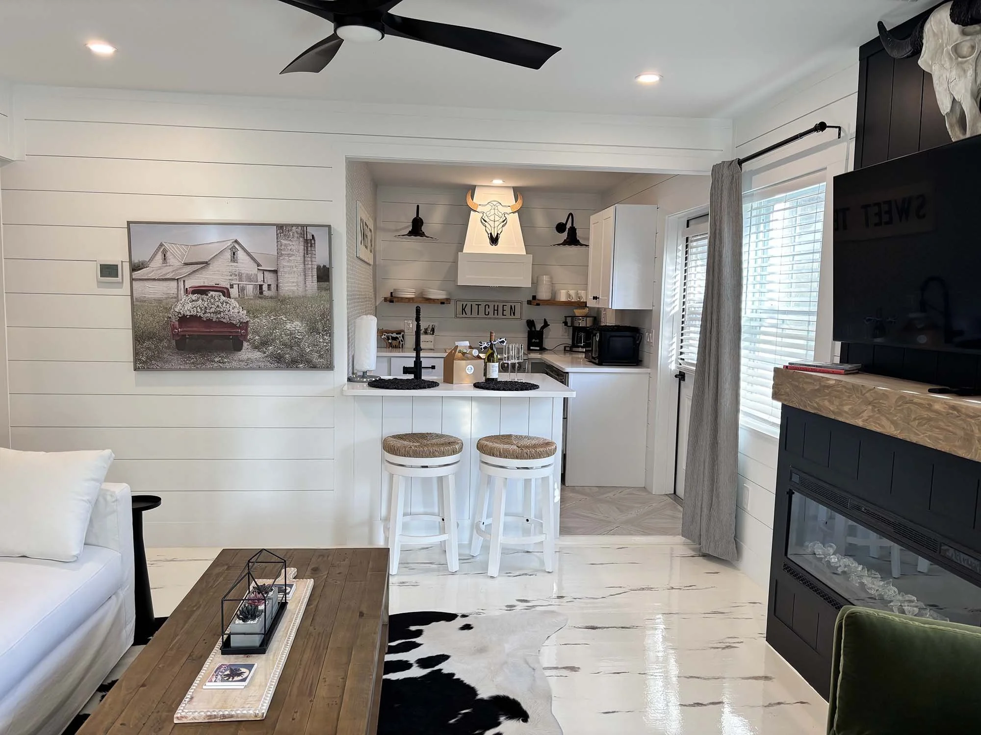 Living room with white shiplap walls, a large farmhouse-style photograph, a fireplace with a black frame, a mounted TV, a wooden coffee table, a black and white cowhide rug, and a kitchen area with a white island, two barstools, and modern decor.