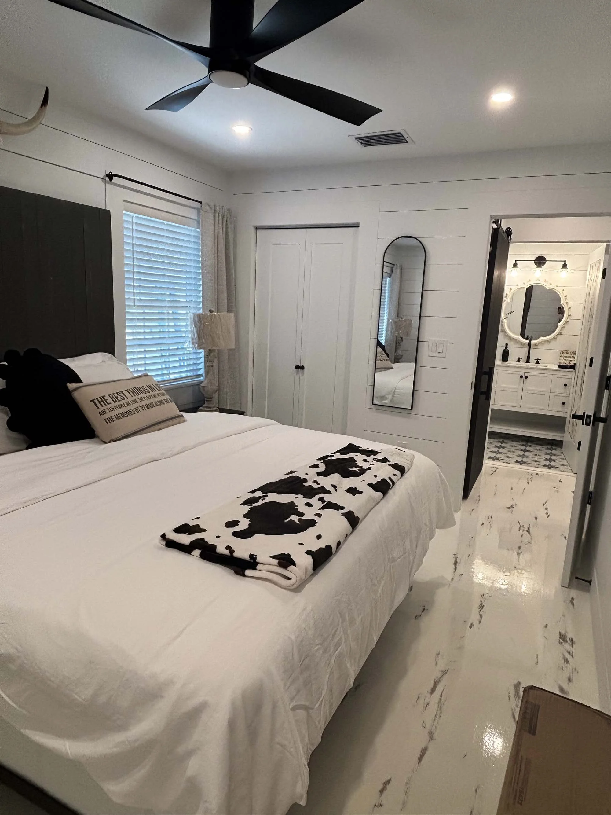 A neatly made bedroom with a white bed, cow print blanket, black and beige pillows, a window with blinds on the left, and a closet with sliding doors. There is a tall mirror on the wall and an en-suite bathroom with a white vanity, round mirror, and 